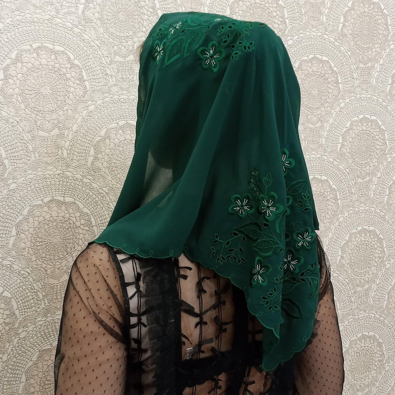 May include: A dark green head covering with floral embroidery. The sheer fabric drapes over the head and shoulders, with the embroidered flowers adding a decorative touch. The edges are scalloped.