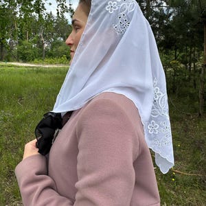 May include: A white, sheer head covering with floral embroidery along the edges. The head covering is draped over a person's head and shoulders, and is paired with a pink coat. The background features green grass and trees.