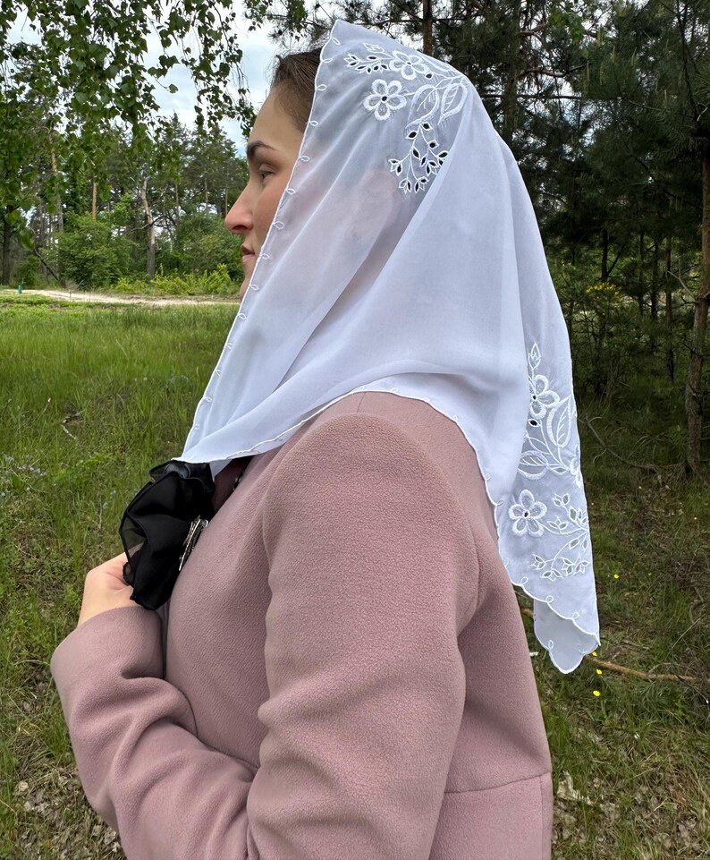 May include: A white, sheer head covering with floral embroidery along the edges. The head covering is draped over a person's head and shoulders, and is paired with a pink coat. The background features green grass and trees.
