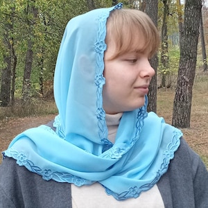 以下が含まれることがあります： 花柄の刺繍が施された水色のヘッドカバー。スカーフは頭と肩にかけられ、花の模様は濃い青色です。背景には木々のある森が写っています。