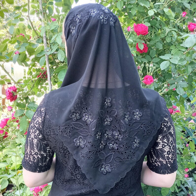 May include: Black head covering with floral embroidery. The head covering is made of a sheer fabric and features a detailed floral design around the edges and on the back. The sleeves of the garment underneath are made of lace.