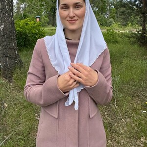 May include: A light pink coat with a matching scarf and a white head covering. The coat has a classic design with a high collar and pockets. The white head covering has a scalloped edge.