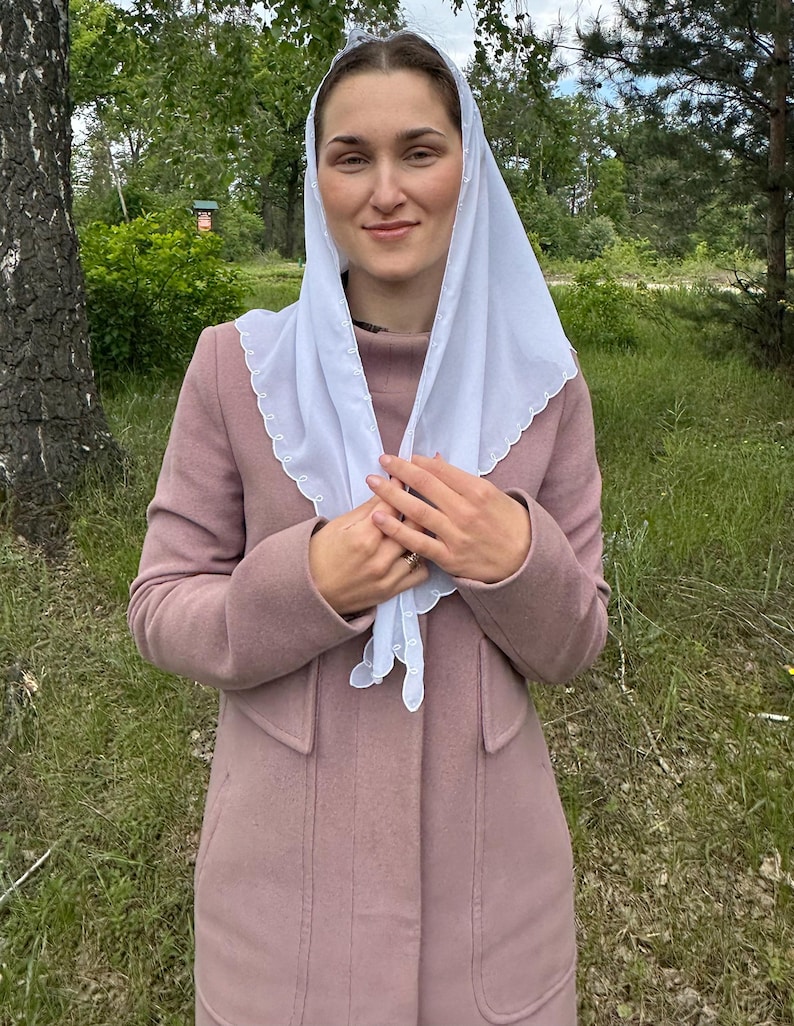 May include: A light pink coat with a matching scarf and a white head covering. The coat has a classic design with a high collar and pockets. The white head covering has a scalloped edge.
