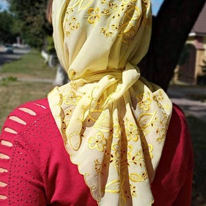 May include: A light yellow headscarf with intricate floral embroidery in gold thread. The scarf is draped over a person's head and shoulders, with the design visible on the edges. The person is wearing a red sweater.