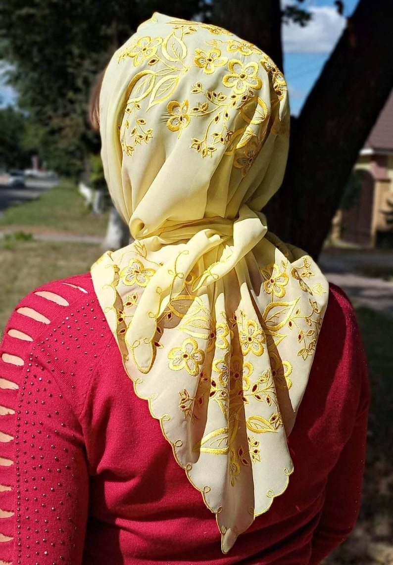 May include: A light yellow headscarf with intricate floral embroidery in gold thread. The scarf is draped over a person's head and shoulders, with the design visible on the edges. The person is wearing a red sweater.