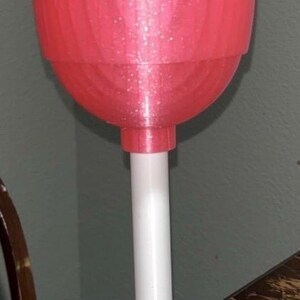 May include: A large, pink, 3D-printed lollipop prop with a white stem and base. The lollipop head has a textured surface and is mounted on a white cylindrical stem. The base is round and white.
