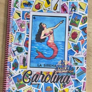May include: Spiral-bound notebook with a vibrant Loteria card design. The central image is a mermaid, with the text "LA SIRENA". The name "Carolina" is printed in cursive. The notebook has a pink spiral binding.