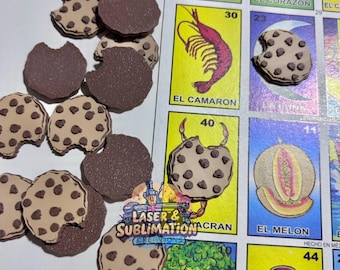 Puede incluir: Una colección de adornos con forma de galleta marrón y beige, algunos con un mordisco, junto a coloridas cartas de Lotería. Las cartas presentan ilustraciones y texto en español. La imagen también incluye el texto "LASER & SUBLIMATION CREATIONS".