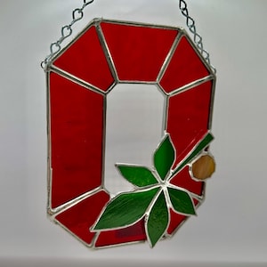 May include: A stained glass decoration in the shape of a red letter "O" with a green leaf and a small tan circle. The glass is outlined with silver-colored metal and hangs from a silver chain.