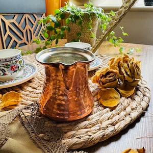 May include: A hammered copper Turkish coffee pot with a long handle, sitting on a woven straw placemat with dried flower petals. The pot is next to a small white teacup and saucer with a floral design.
