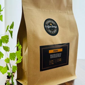 May include: A brown paper bag of coffee beans with the text "Coffee Denya" and "PERU" on the label. The label includes information about the coffee's flavour profile. Coffee beans are scattered on the wooden surface.