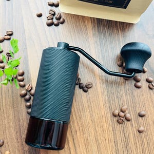 Op de afbeelding: Een zwarte handmatige koffiemolen met een getextureerde behuizing en een donkerhouten handvat. Koffiebonen en groene takjes liggen verspreid op een houten oppervlak, met een zak koffiebonen op de achtergrond. De molen is ontworpen voor handmatig gebruik.