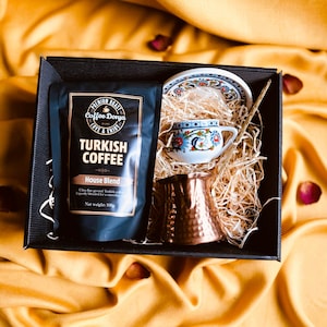 May include: A black gift box filled with Turkish coffee, a copper cezve, a small white and blue patterned cup and saucer, and a wooden stirrer. The box is sitting on a yellow fabric surface.