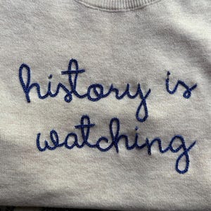 K&ouml;nnte beinhalten: Ein beigefarbenes Sweatshirt mit Rundhalsausschnitt und dem dunkelblau gestickten Text "history is watching" auf der Brust. Das Kleidungsst&uuml;ck hat lange &Auml;rmel und einen gerippten Halsausschnitt mit einer kursiven Schrift.