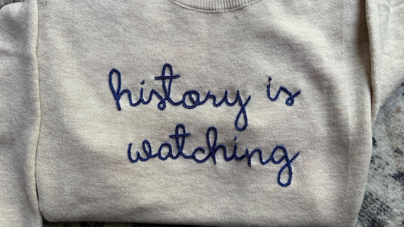 K&ouml;nnte beinhalten: Ein beigefarbenes Sweatshirt mit Rundhalsausschnitt und dem dunkelblau gestickten Text "history is watching" auf der Brust. Das Kleidungsst&uuml;ck hat lange &Auml;rmel und einen gerippten Halsausschnitt mit einer kursiven Schrift.