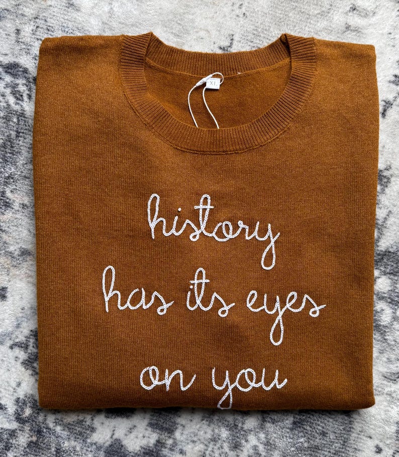 K&ouml;nnte beinhalten: Ein tabakbrauner Pullover mit Rundhalsausschnitt und wei&szlig;em, in Schreibschrift gesticktem Text, der history has its eyes on you lautet. Das Kleidungsst&uuml;ck hat ein wei&szlig;es XL-Gr&ouml;&szlig;enetikett, das mit einem d&uuml;nnen Kunststoffband am Kragen befestigt ist.