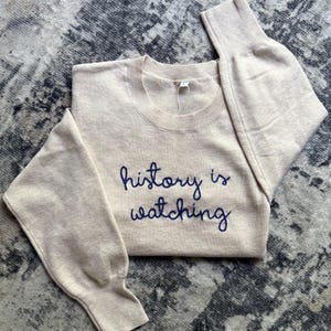 K&ouml;nnte beinhalten: Ein beigefarbener Langarmpullover mit Rundhalsausschnitt und dem dunkelblau gestickten Text "history is watching" auf der Brust. Das Kleidungsst&uuml;ck hat gerippte B&uuml;ndchen und einen gerippten Halsausschnitt sowie ein wei&szlig;es Etikett.