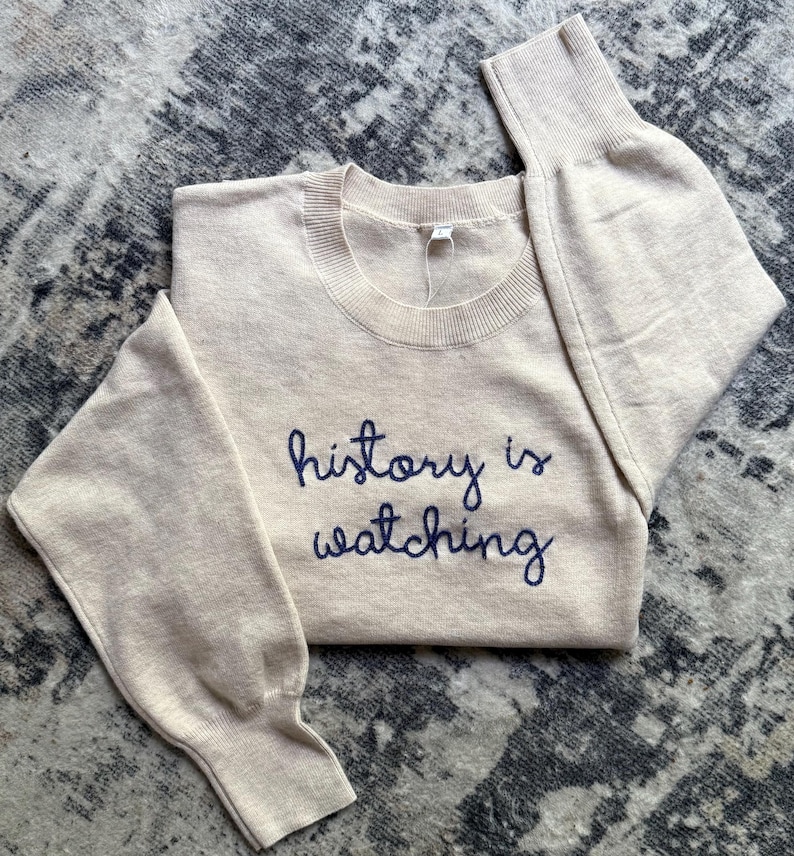 K&ouml;nnte beinhalten: Ein beigefarbener Langarmpullover mit Rundhalsausschnitt und dem dunkelblau gestickten Text "history is watching" auf der Brust. Das Kleidungsst&uuml;ck hat gerippte B&uuml;ndchen und einen gerippten Halsausschnitt sowie ein wei&szlig;es Etikett.