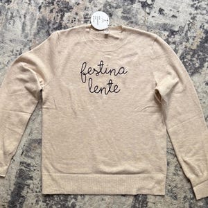 K&ouml;nnte beinhalten: Ein beigefarbener Langarmpullover mit Rundhalsausschnitt und dem schwarzen Sticktext "festina lente" mittig auf der Brust. Das Kleidungsst&uuml;ck hat einen gerippten Halsausschnitt, &Auml;rmelb&uuml;ndchen und Saum sowie ein kleines wei&szlig;es Etikett am Kragen.