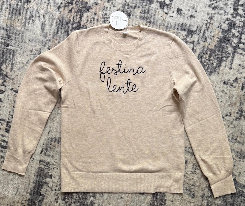 K&ouml;nnte beinhalten: Ein beigefarbener Langarmpullover mit Rundhalsausschnitt und dem schwarzen Sticktext "festina lente" mittig auf der Brust. Das Kleidungsst&uuml;ck hat einen gerippten Halsausschnitt, &Auml;rmelb&uuml;ndchen und Saum sowie ein kleines wei&szlig;es Etikett am Kragen.