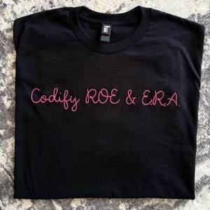 May include: Black t-shirt with the pink text "Codify ROE & ERA." The shirt is made of a soft material and has a crew neck. The text is in a cursive font.