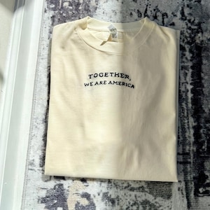 May include: Cream-colored t-shirt with the embroidered text "TOGETHER, WE ARE AMERICA" in black. The shirt is folded neatly and appears to be made of a soft material. The background is a patterned rug.