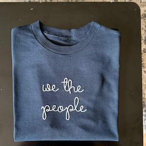 May include: A dark navy blue short-sleeved t-shirt featuring the text "we the people" embroidered in white thread across the center of the chest. The shirt has a classic crew neckline and is folded neatly to display the front graphic clearly.