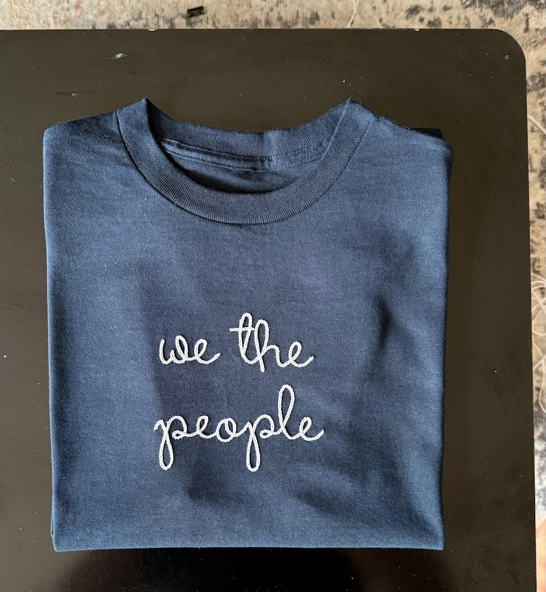 May include: A dark navy blue short-sleeved t-shirt featuring the text "we the people" embroidered in white thread across the center of the chest. The shirt has a classic crew neckline and is folded neatly to display the front graphic clearly.