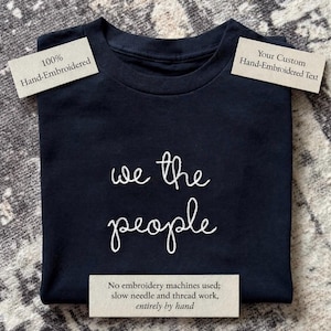 May include: A black t-shirt featuring white hand-embroidered text that reads "we the people" in a cursive script. The shirt includes labels stating "100% Hand-Embroidered," "Your Custom Hand-Embroidered Text," and "No embroidery machines used."