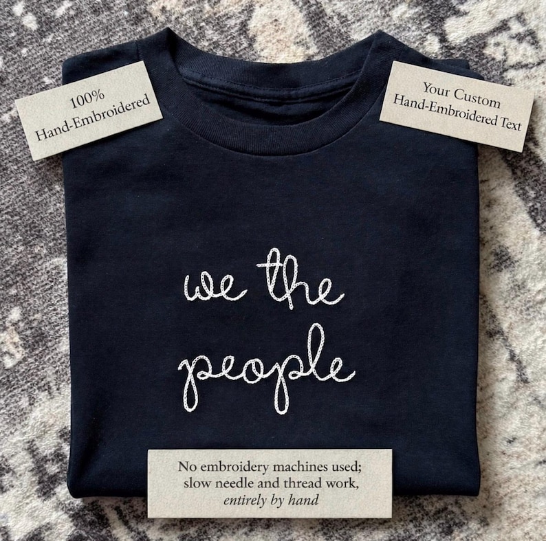 May include: A black t-shirt featuring white hand-embroidered text that reads "we the people" in a cursive script. The shirt includes labels stating "100% Hand-Embroidered," "Your Custom Hand-Embroidered Text," and "No embroidery machines used."