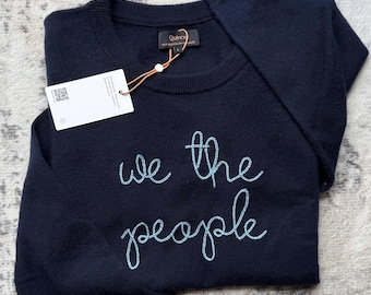 Könnte beinhalten: Ein dunkelblauer Langarmpullover mit dem Schriftzug we the people, der in hellblauem Garn auf die Brust gestickt ist. Das Kleidungsstück hat ein Quince-Markenetikett am Halsausschnitt und ein weißes, rechteckiges Produktetikett an einer Schnur.