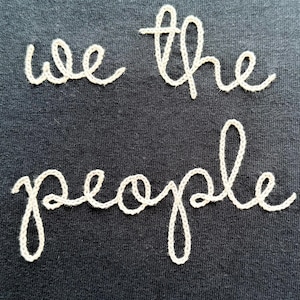 May include: A black fabric surface featuring the text we the people embroidered in a light beige thread. The lettering is rendered in a cursive script style, with the words arranged in two lines, centered against the dark background.
