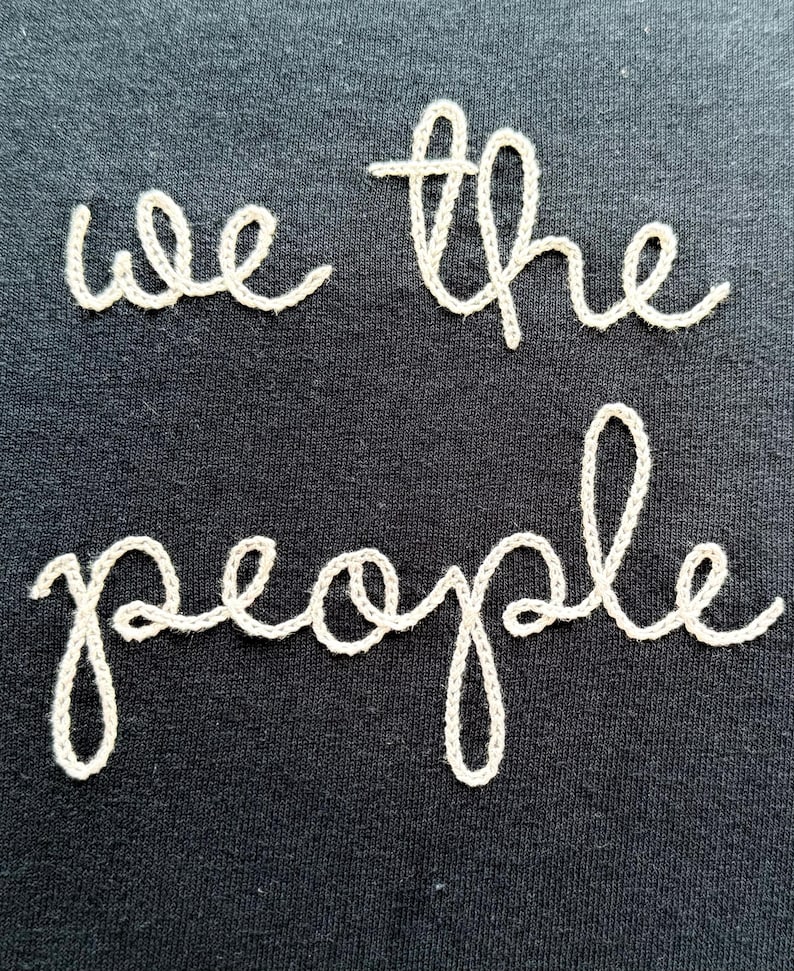 May include: A black fabric surface featuring the text we the people embroidered in a light beige thread. The lettering is rendered in a cursive script style, with the words arranged in two lines, centered against the dark background.