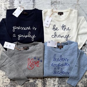 May include: Four long-sleeved crewneck sweaters in navy, cream, gray, and light blue. The navy sweater reads "pressure is a privilege," the cream reads "be the change," the gray features a peace sign and stars, and the light blue reads "leaders empower leaders."