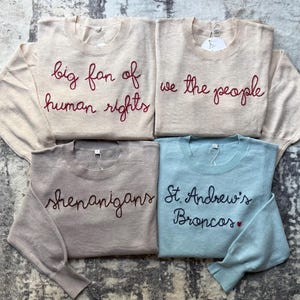 K&ouml;nnte beinhalten: Vier lang&auml;rmelige Pullover mit Rundhalsausschnitt in Beige, Hellbraun und Hellblau. Der obere linke Pullover zeigt den roten Schriftzug big fan of human rights. Oben rechts steht we the people. Unten links steht shenanigans und unten rechts St. Andrew's Broncos mit einem kleinen roten Herz.