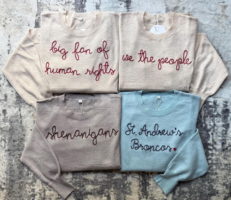 K&ouml;nnte beinhalten: Vier lang&auml;rmelige Pullover mit Rundhalsausschnitt in Beige, Hellbraun und Hellblau. Der obere linke Pullover zeigt den roten Schriftzug big fan of human rights. Oben rechts steht we the people. Unten links steht shenanigans und unten rechts St. Andrew's Broncos mit einem kleinen roten Herz.