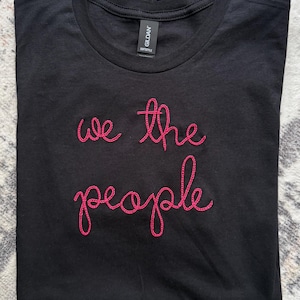 May include: A black short-sleeved t-shirt featuring the text "we the people" embroidered in bright pink thread across the chest. The garment has a crew neckline and a Gildan Softstyle brand label visible on the inside of the collar.