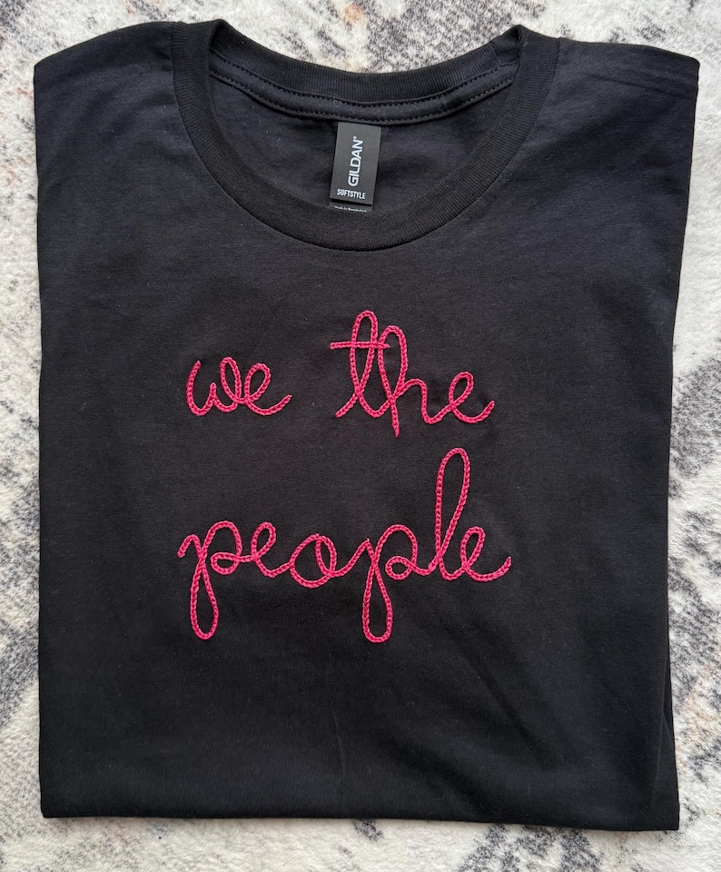May include: A black short-sleeved t-shirt featuring the text "we the people" embroidered in bright pink thread across the chest. The garment has a crew neckline and a Gildan Softstyle brand label visible on the inside of the collar.
