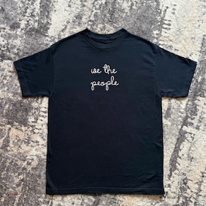 May include: A black short-sleeved t-shirt featuring the text "we the people" written in white cursive script across the center of the chest. The garment has a crew neckline and a standard fit, showcasing a clean and simple design on a solid black fabric.