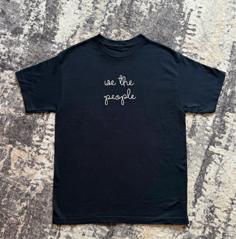 May include: A black short-sleeved t-shirt featuring the text "we the people" written in white cursive script across the center of the chest. The garment has a crew neckline and a standard fit, showcasing a clean and simple design on a solid black fabric.