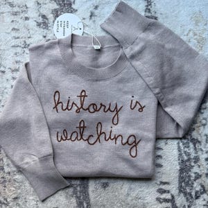 K&ouml;nnte beinhalten: Ein beigefarbener Langarm-Strickpullover mit geripptem Rundhalsausschnitt und B&uuml;ndchen. Auf der Vorderseite befindet sich der dunkelbraun gestickte Text "history is watching". Ein rundes wei&szlig;es Etikett am Kragen tr&auml;gt die Aufschrift "KEEP IN FASHION STAY IN STYLE".