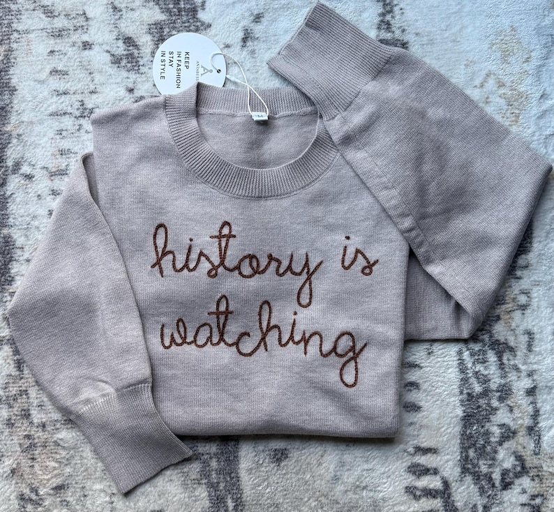 K&ouml;nnte beinhalten: Ein beigefarbener Langarm-Strickpullover mit geripptem Rundhalsausschnitt und B&uuml;ndchen. Auf der Vorderseite befindet sich der dunkelbraun gestickte Text "history is watching". Ein rundes wei&szlig;es Etikett am Kragen tr&auml;gt die Aufschrift "KEEP IN FASHION STAY IN STYLE".