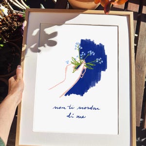 May include: Framed art print featuring a hand holding a sprig of blue forget-me-nots against a blue background. The artwork includes the Italian phrase "non ti scordar di me" in dark blue script, set within a white mat and a light wood frame.