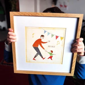Puede incluir: Una impresión enmarcada con una conmovedora ilustración de un padre y un niño bailando. El padre lleva una camisa naranja y pantalones grises, sosteniendo la mano del niño. El niño lleva una camisa verde y pantalones rojos. Se lee "PAPA BALLO" debajo.