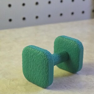 May include: A small, green dumbbell with a textured surface. The dumbbell has a rectangular weight on each end, connected by a cylindrical handle. The background is a light-colored surface with a pegboard.