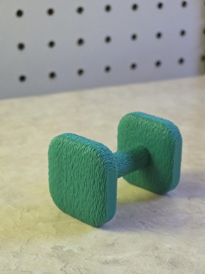 May include: A small, green dumbbell with a textured surface. The dumbbell has a rectangular weight on each end, connected by a cylindrical handle. The background is a light-colored surface with a pegboard.