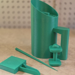May include: A collection of teal 3D-printed objects, including a cylindrical container with a handle, a small stand, a flat square with a handle, and a long, thin rod. The objects are arranged on a light brown surface.