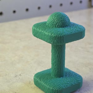 May include: A textured, teal-colored sculpture with a unique design. It features a square base, a cylindrical stem, and a square top with a rounded, textured sphere. The piece is set against a neutral background.