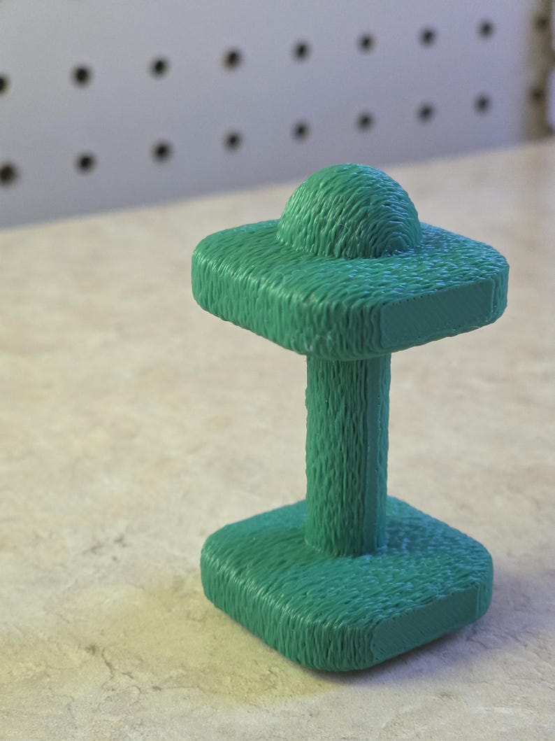 May include: A textured, teal-colored sculpture with a unique design. It features a square base, a cylindrical stem, and a square top with a rounded, textured sphere. The piece is set against a neutral background.