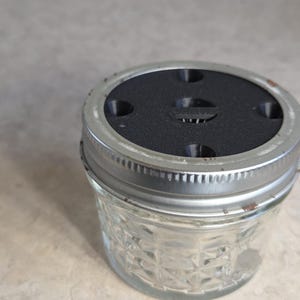 May include: A clear glass jar with a decorative diamond pattern, topped with a black lid and a silver metal rim. The lid has several small holes. The jar is sitting on a light-colored surface.